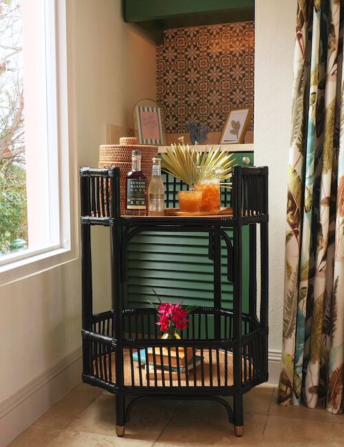 A small wooden bar cart with drinks, a vase, and decor is positioned near a window and patterned curtain.