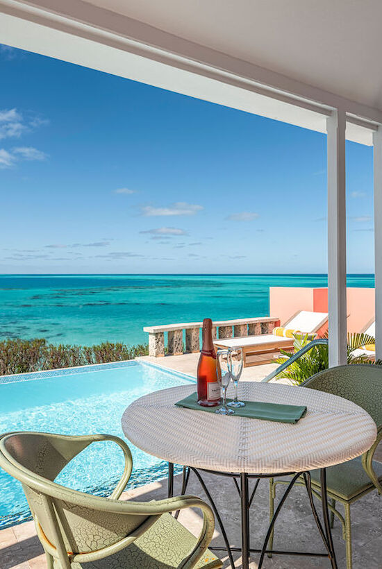 A coastal patio with a table, chairs, wine, and ocean view. An infinity pool and clear skies complete the serene scene.