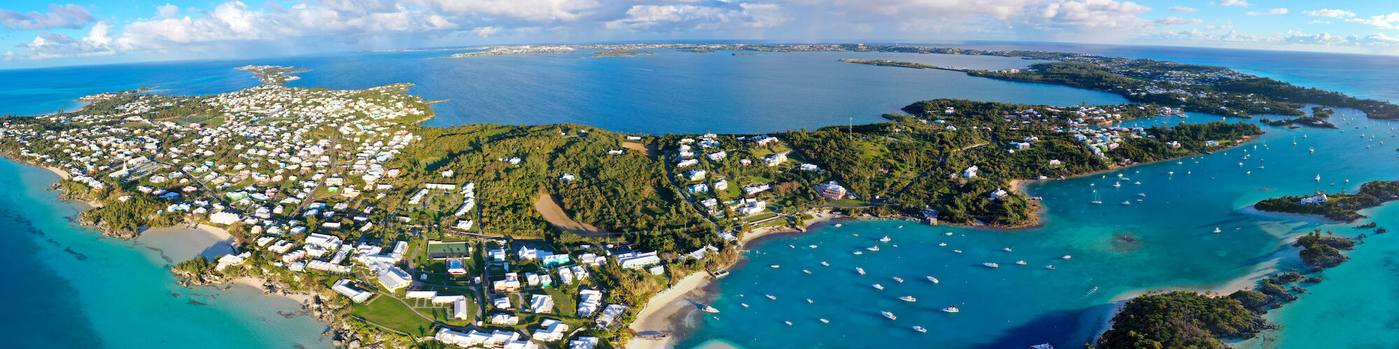 Aerial view of a coastal area with lush greenery, houses, and turquoise waters with boats, under a blue sky with scattered clouds.