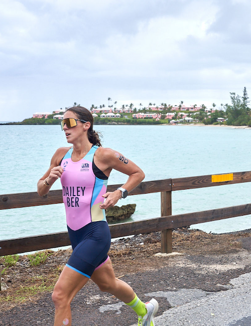 A person is running along a waterfront road, wearing athletic gear and sunglasses, with water and trees in the background.