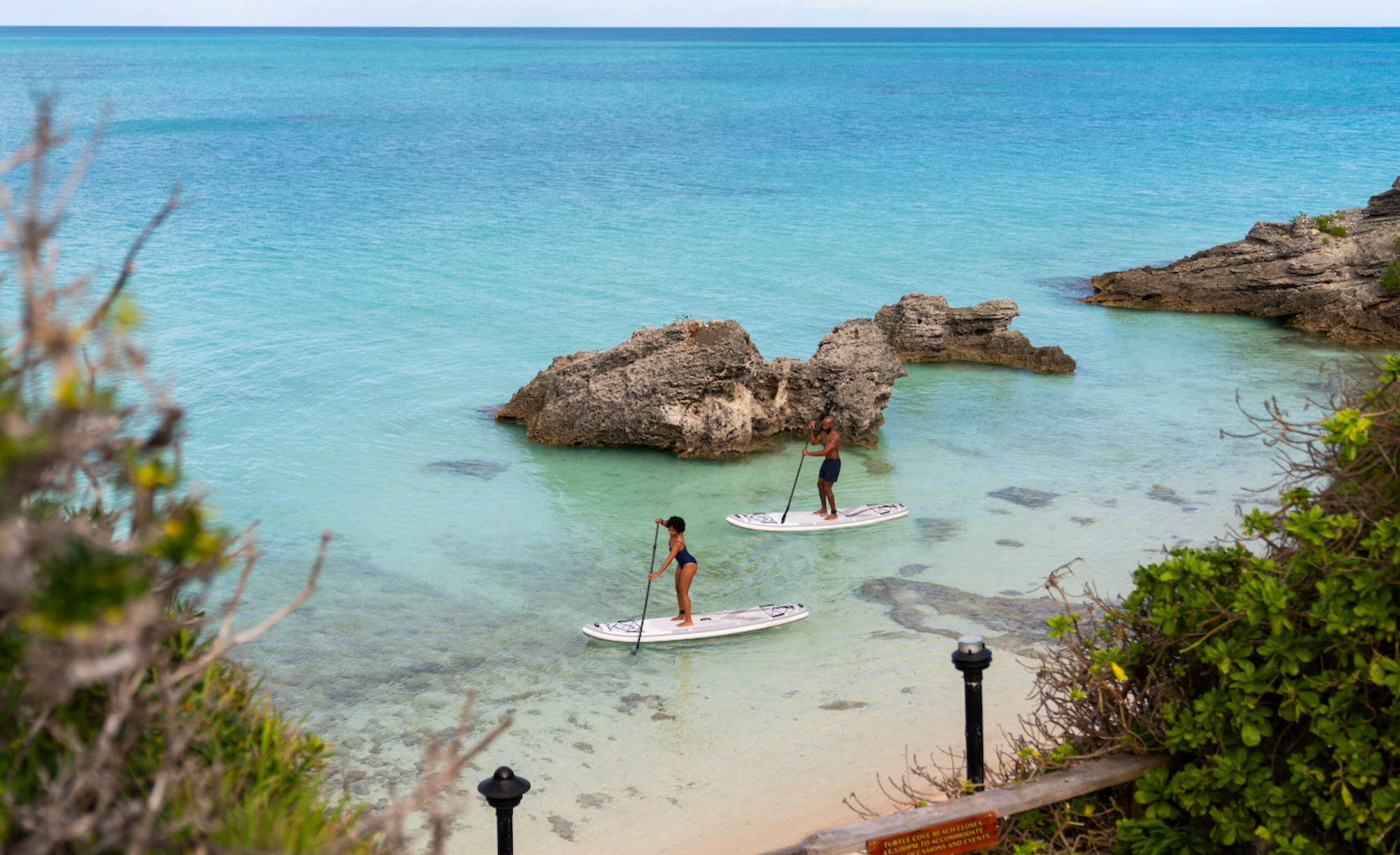 Two people paddleboarding on clear turquoise water near rocks, with greenery framing the scene.