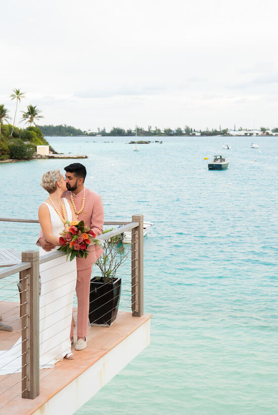 A couple stands on a deck overlooking a serene body of water with boats, surrounded by lush greenery, holding a bouquet.