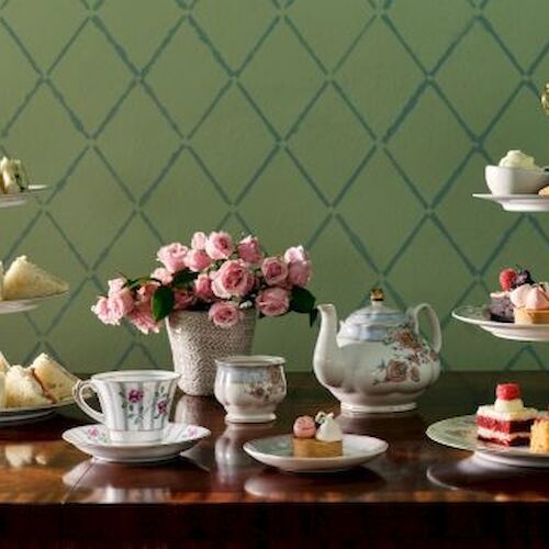 An elegant tea setup with tiered trays of sandwiches and desserts, teacups, a teapot, and pink flowers on a wooden table.