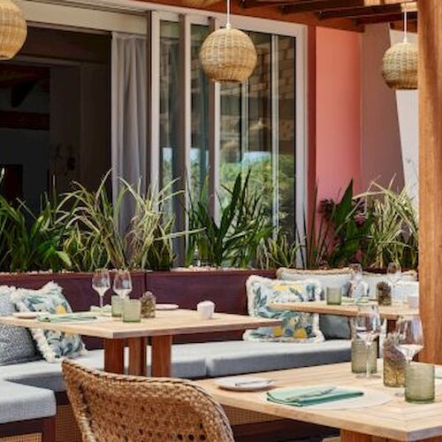 The image shows an outdoor restaurant seating area with wooden tables, cushioned benches, wicker chairs, and hanging lamps.