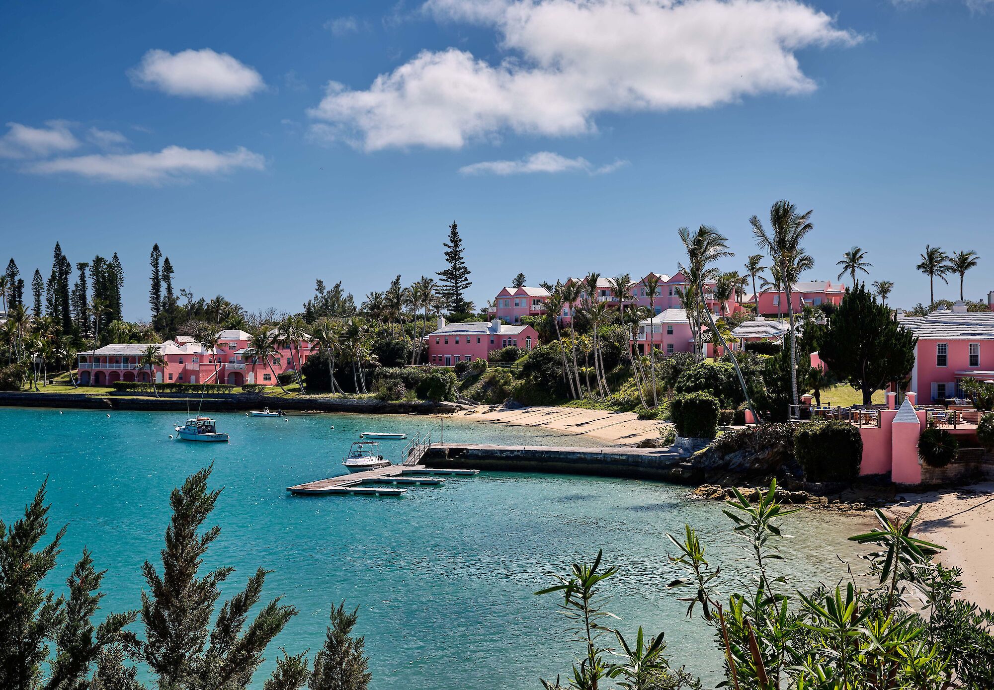 Coastal scene with pink buildings, turquoise water, palm trees, and boats, under a clear blue sky.