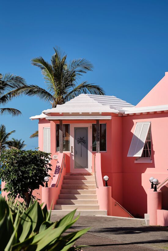 A pink house with white shutters is set against a backdrop of palm trees and a clear blue sky, overlooking the ocean.