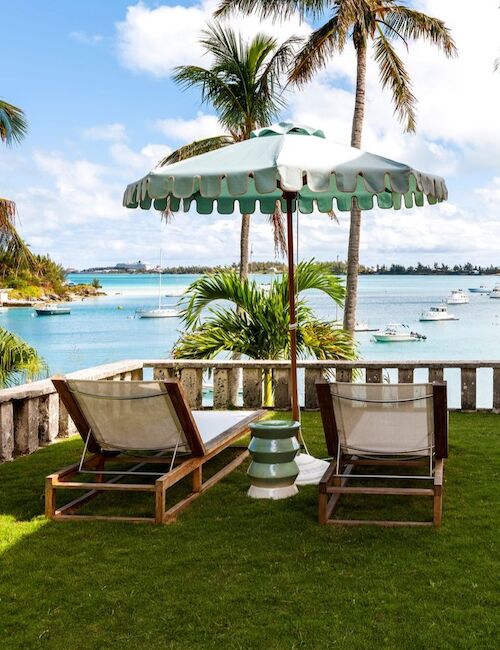 Two lounge chairs and an umbrella on a grassy area overlooking a tropical bay with palm trees and boats under a blue sky.