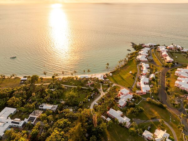 An aerial view of a coastline with a sunlit sea, a curved road of white-roofed houses, and lush green trees along the shore, peaceful and sunny.