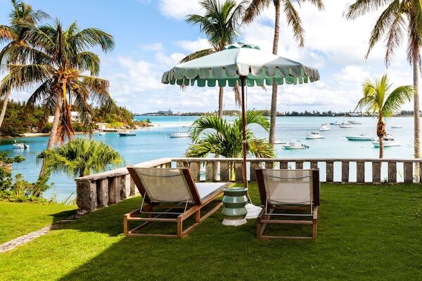 Two lounge chairs and an umbrella sit on a grassy area overlooking a tropical beach with palm trees and boats on the water.