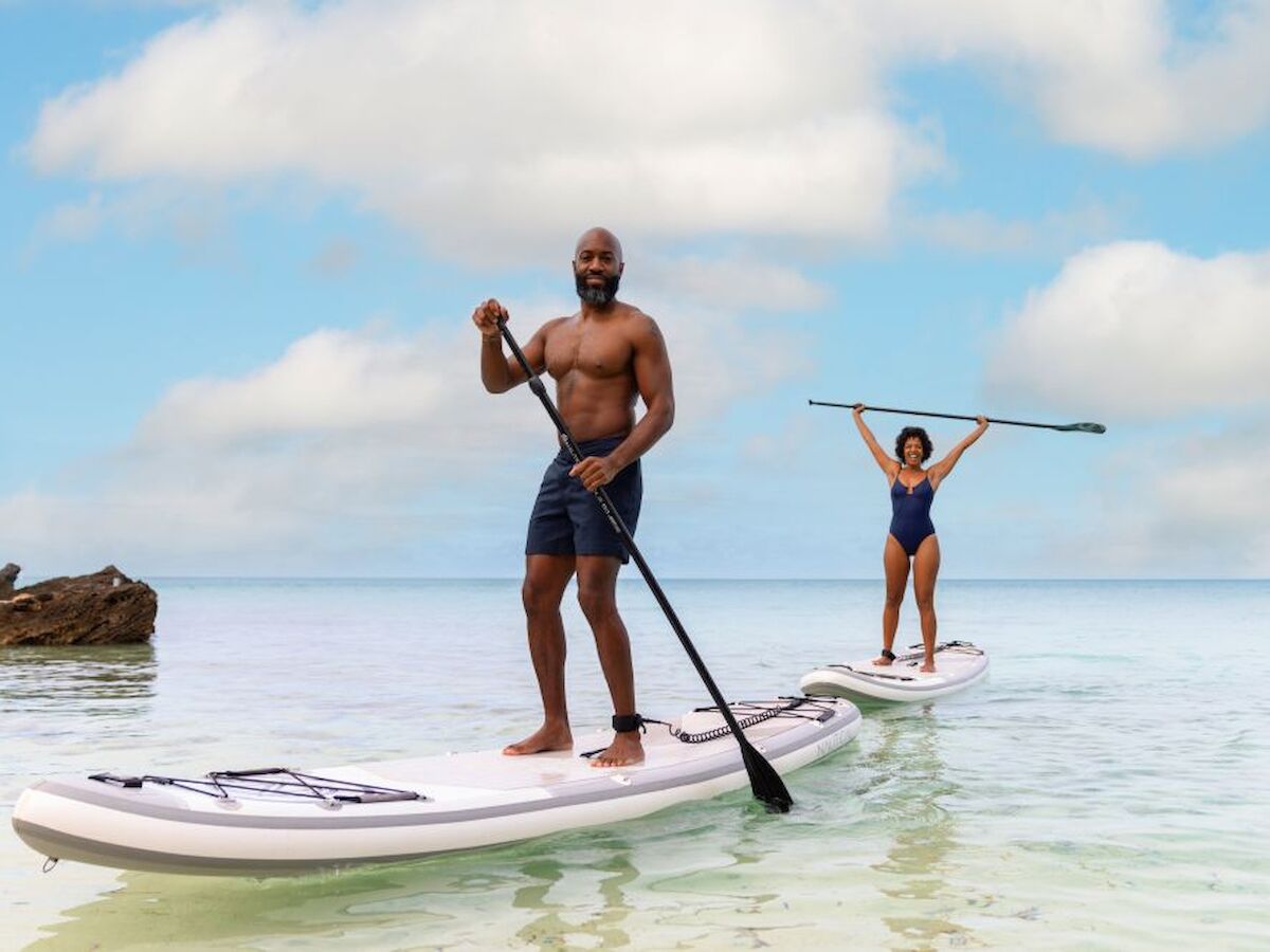Two people are stand-up paddleboarding on a calm, clear sea under a partly cloudy sky.