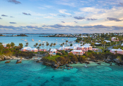 A scenic coastal view with turquoise waters, pink houses, and boats on the horizon under a partly cloudy sky.