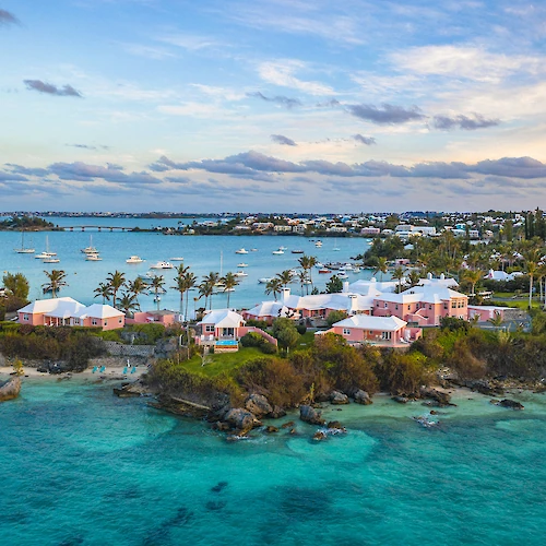 A scenic coastal view with turquoise waters, pink houses, and boats on the horizon under a partly cloudy sky.