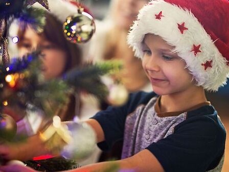 A child wearing a Santa hat decorates a Christmas tree, with others in the background, creating a festive holiday scene.