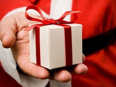 A person in a Santa suit holds a small gift box with a red ribbon in their hand.