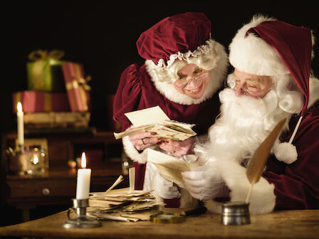 Two people dressed as Santa and Mrs. Claus sort through letters and money at a cozy desk, candles flicker, gifts pile in the background.