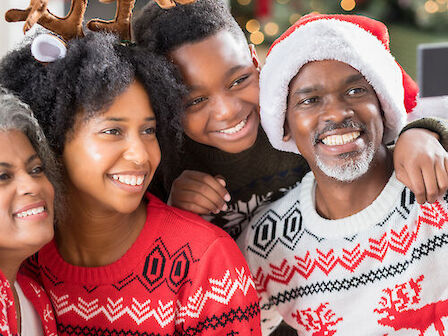 A family wearing festive sweaters and hats, with Christmas decorations in the background, smiles happily for a holiday photo.