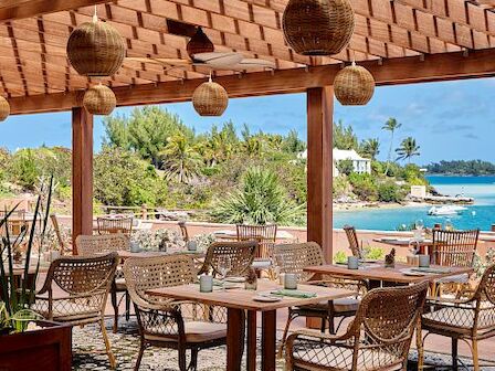 An outdoor dining area with wicker chairs, wooden tables, and pendant lights under a pergola, overlooking a scenic coastal view.
