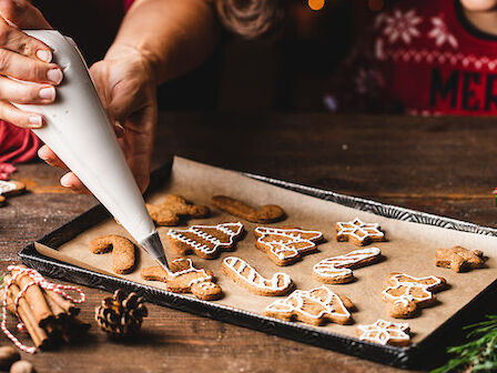 Hands decorating festive cookies with icing on a tray, next to holiday decor on a wooden table.