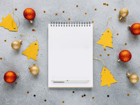 Notebook and pen surrounded by golden and red baubles and yellow tree-shaped tags on a gray surface with scattered confetti.