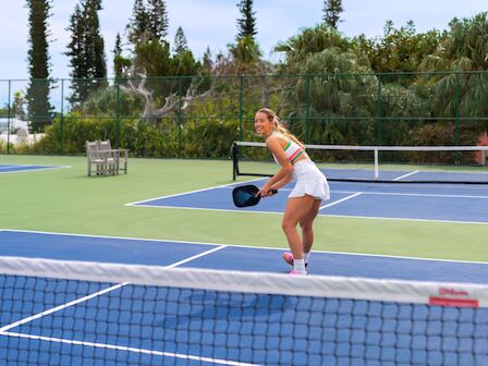 A person is playing pickleball on an outdoor court surrounded by trees, holding a paddle and wearing a white outfit.