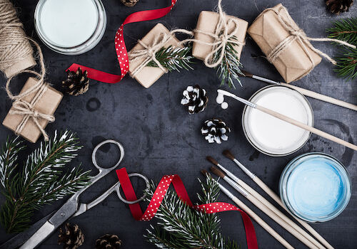 Crafting scene with wrapped gifts, twine, ribbons, pinecones, paintbrushes, paints, and evergreen branches on a dark surface.