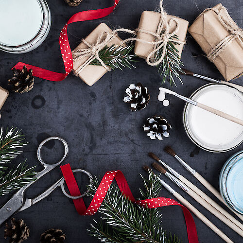 Crafting scene with wrapped gifts, twine, ribbons, pinecones, paintbrushes, paints, and evergreen branches on a dark surface.