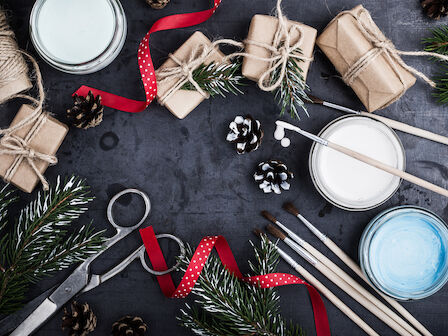 Gift wrapping supplies, pine branches, pinecones, brushes, paints, and scissors are arranged creatively on a dark surface.