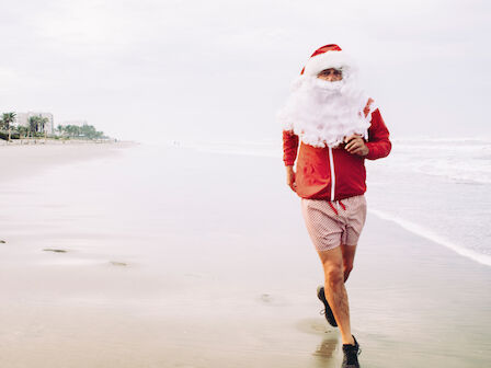 A person in a Santa outfit jogs along a beach, wearing a red jacket, shorts, and black shoes, with a blurred ocean backdrop.