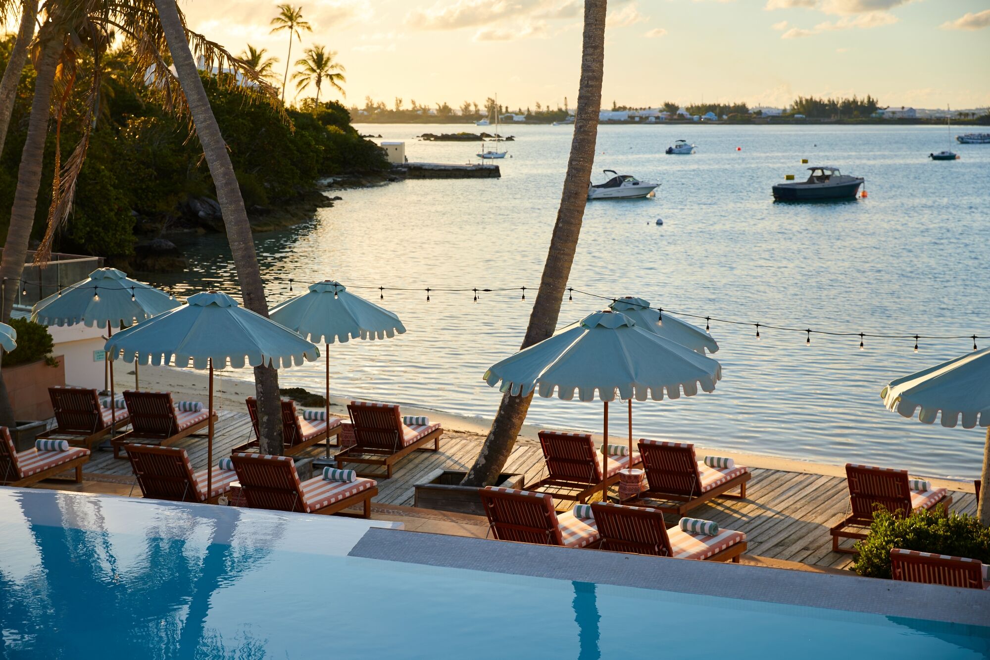 A tropical beachfront scene with palm trees, sun loungers under blue parasols, a calm sea with boats, and a wooden deck by the water.