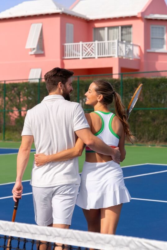A couple stands on a tennis court, embracing after a match, with pink houses in the background.