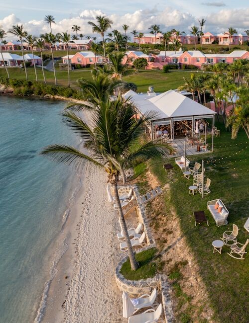 A tropical beachfront resort with palm trees, a sandy shoreline, striped tents, a gazebo, and seaside seating on a grassy lawn.