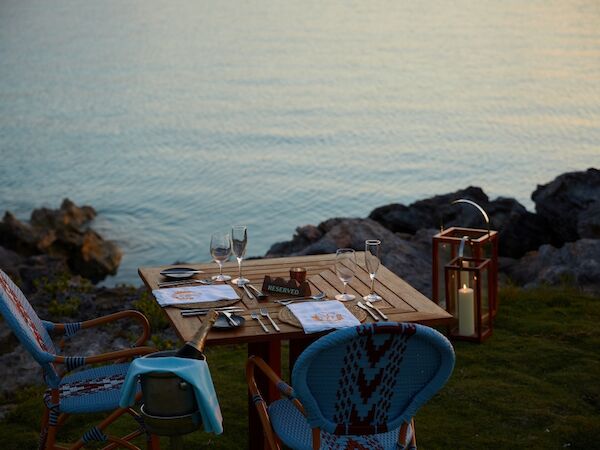 A seaside picnic setup with a wooden table, blue chairs, utensils, plates, and candles by rocky shore at sunset.