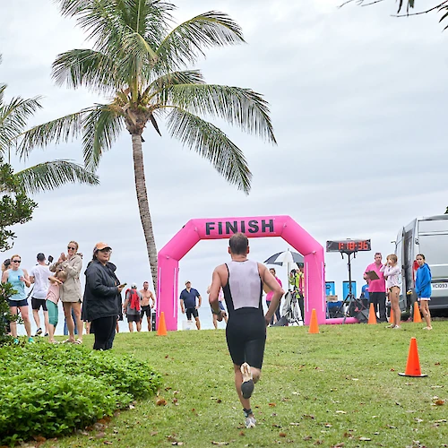 A person is approaching the pink "FINISH" arch at an outdoor event, with spectators and palm trees in the background.