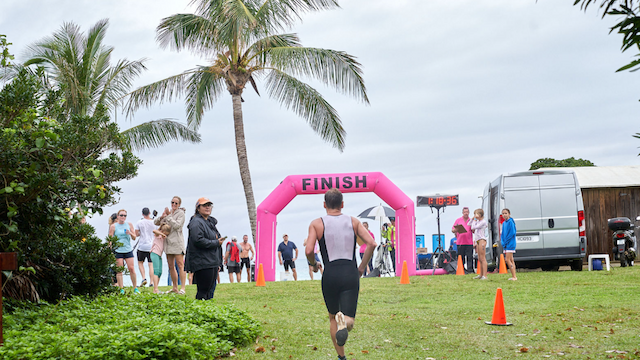 A person is approaching the pink "FINISH" arch at an outdoor event, with spectators and palm trees in the background.