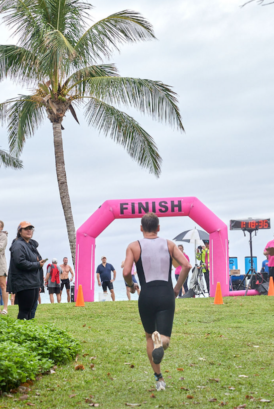 A runner approaches a pink finish line in an outdoor race. Spectators stand nearby, and there's a van parked in the background.