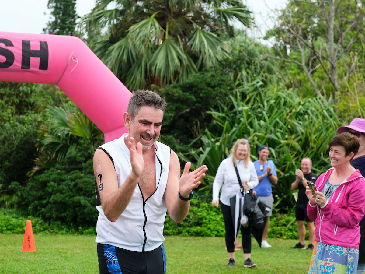 A man is clapping near a pink "FINISH" arch, with onlookers in the background in a lush outdoor setting.