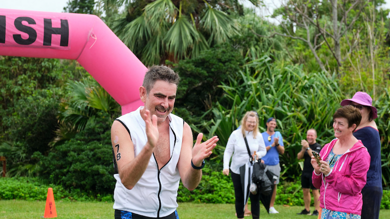 A man is clapping near a pink "FINISH" arch, with onlookers in the background in a lush outdoor setting.