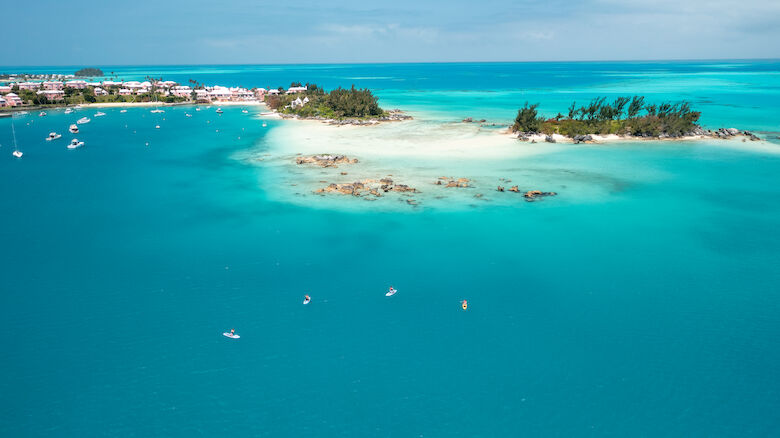 A scenic aerial view of a turquoise ocean with several small islands, boats, and a coastal settlement in the background under a clear sky.