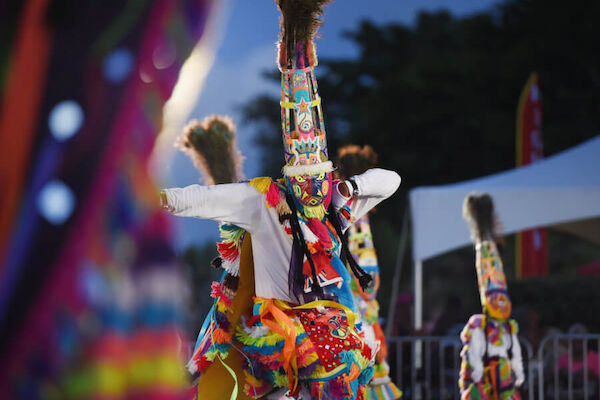 People in colorful, traditional costumes are dancing outdoors, wearing tall, decorative hats, surrounded by a festive atmosphere.
