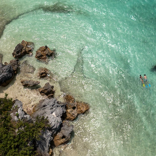 An aerial view of clear turquoise water, rocks, and two people swimming near the shore, surrounded by a natural coastal landscape.