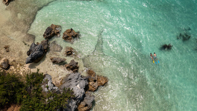An aerial view of clear turquoise water, rocks, and two people swimming near the shore, surrounded by a natural coastal landscape.