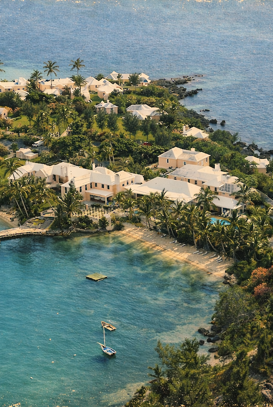 A tropical coastal resort with white villas, palm trees, and a turquoise lagoon, small boats anchored near a curved sandy shore by the blue sea.