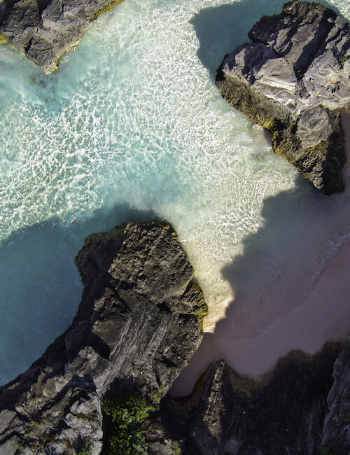 An aerial view of turquoise water and a sandy beach surrounded by dark, rocky formations, possibly a coastal area.