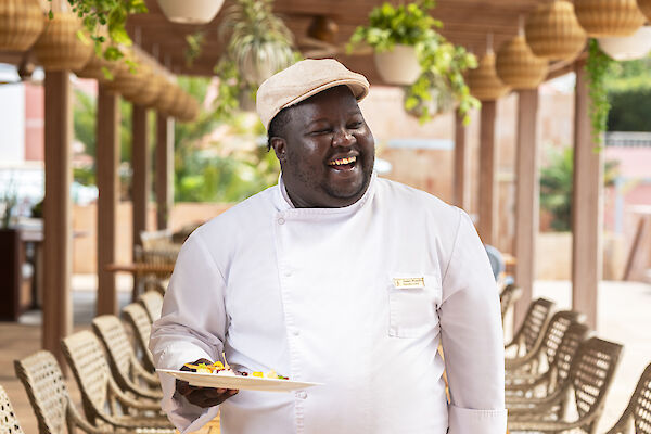 A person in chef attire holds a dish in an outdoor dining area with wooden tables and plants.