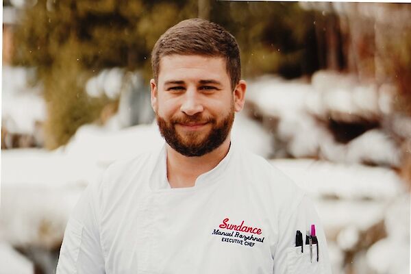 A person in a white chef's coat with pens in the pocket, standing outdoors in a snowy setting, smiling at the camera.