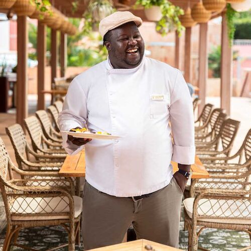 A smiling chef in a white jacket and cap serves food on a plate in a sunny outdoor restaurant patio.