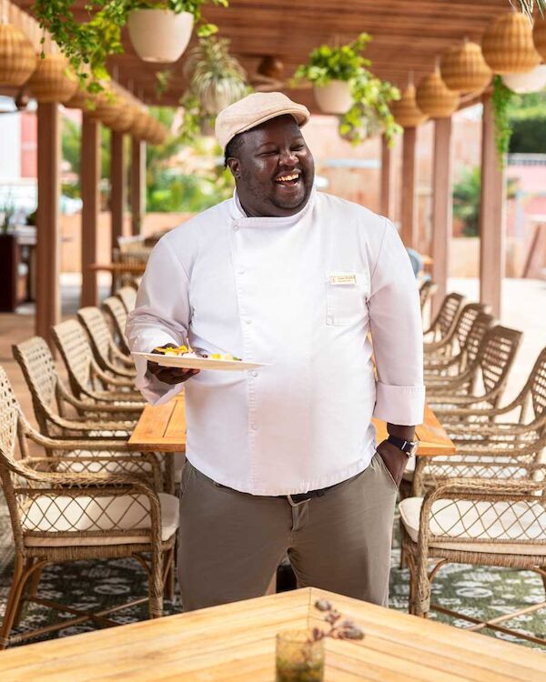 A smiling chef in a white jacket and cap serves food on a plate in a sunny outdoor restaurant patio.
