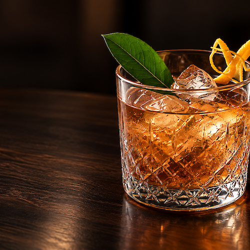 Amber whisky cocktail with ice in a faceted glass, garnished with a green leaf and orange peel twist, on a wooden bar.