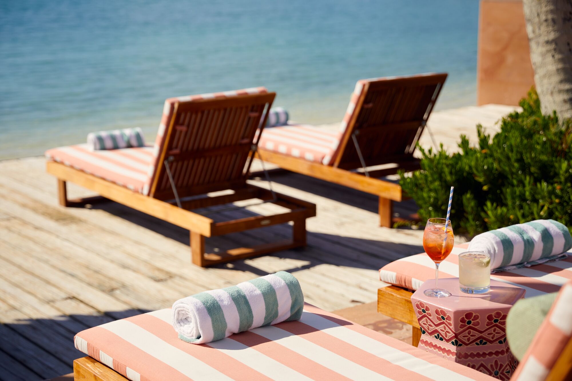 Beach scene with sun loungers, striped towels, drinks on a wooden deck, overlooking a tranquil sea under clear skies.