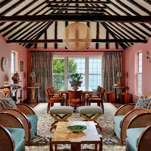 A cozy living room with pink walls, beamed ceiling, multiple chairs, a table, colorful decor, and a large window.